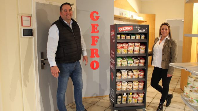Auf diesem Bild sehen Sie einen Mann und eine Frau die im GEFRO-Laden in Memmingen. Sie stehen beide neben einem Produkt-Aufsteller in dem viele verschiedene GEFRO-Produkte entahlten sind.