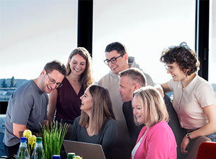 Sechs Personen arbeiten gemeinsam an einem Laptop im Büro, Teammeeting in heller Arbeitsumgebung