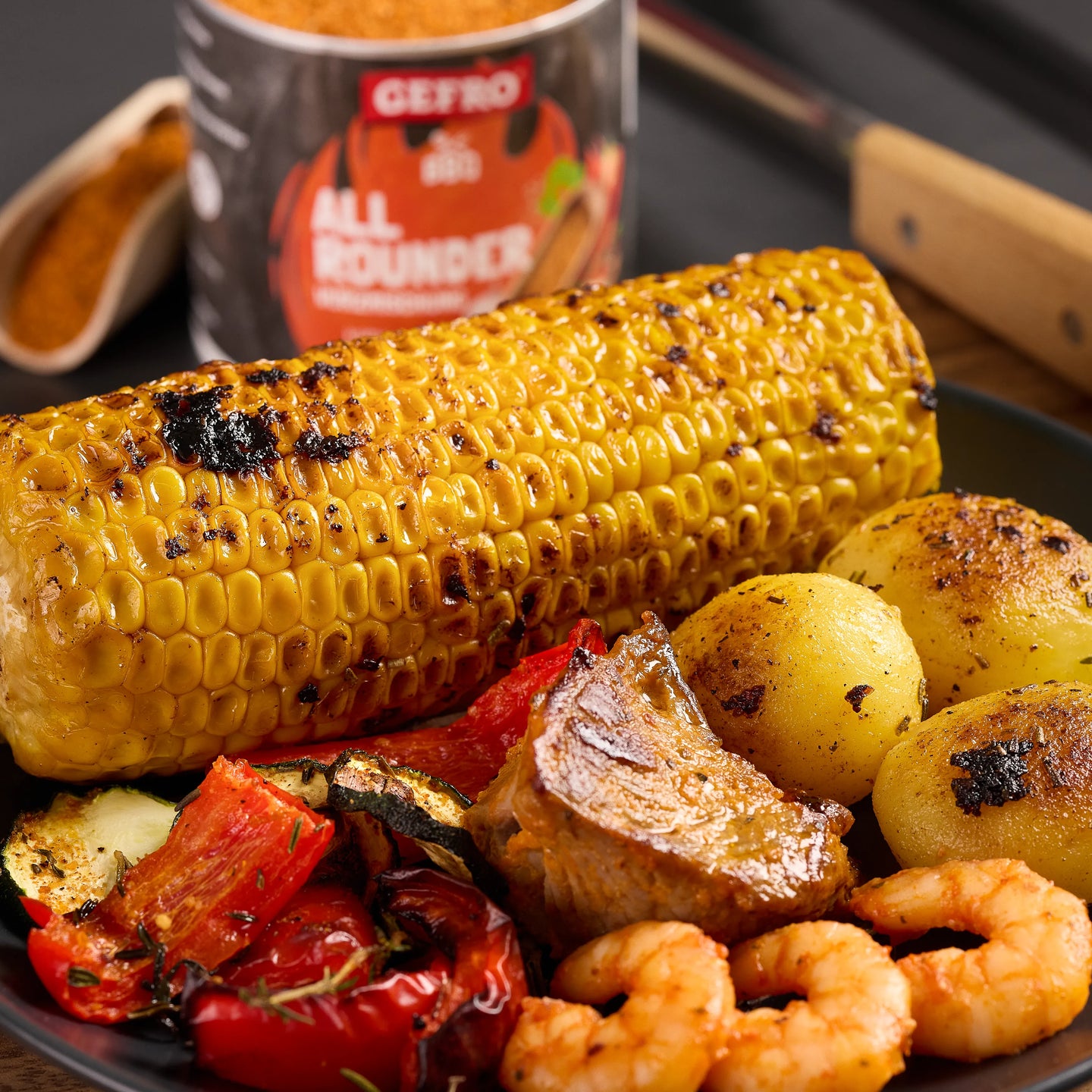Gegrillter Maiskolben mit Kartoffeln, Garnelen, Fleischstück und Gemüse auf einem Teller, Gewürzdose im Hintergrund