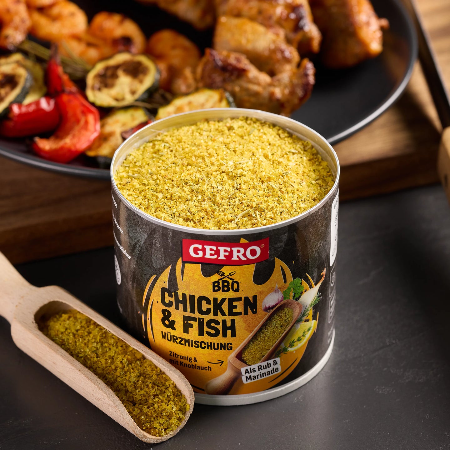 Offene Dose GEFRO BBQ Chicken & Fish Würzmischung mit Holzschaufel, vor gegrilltem Hähnchen und Gemüse.