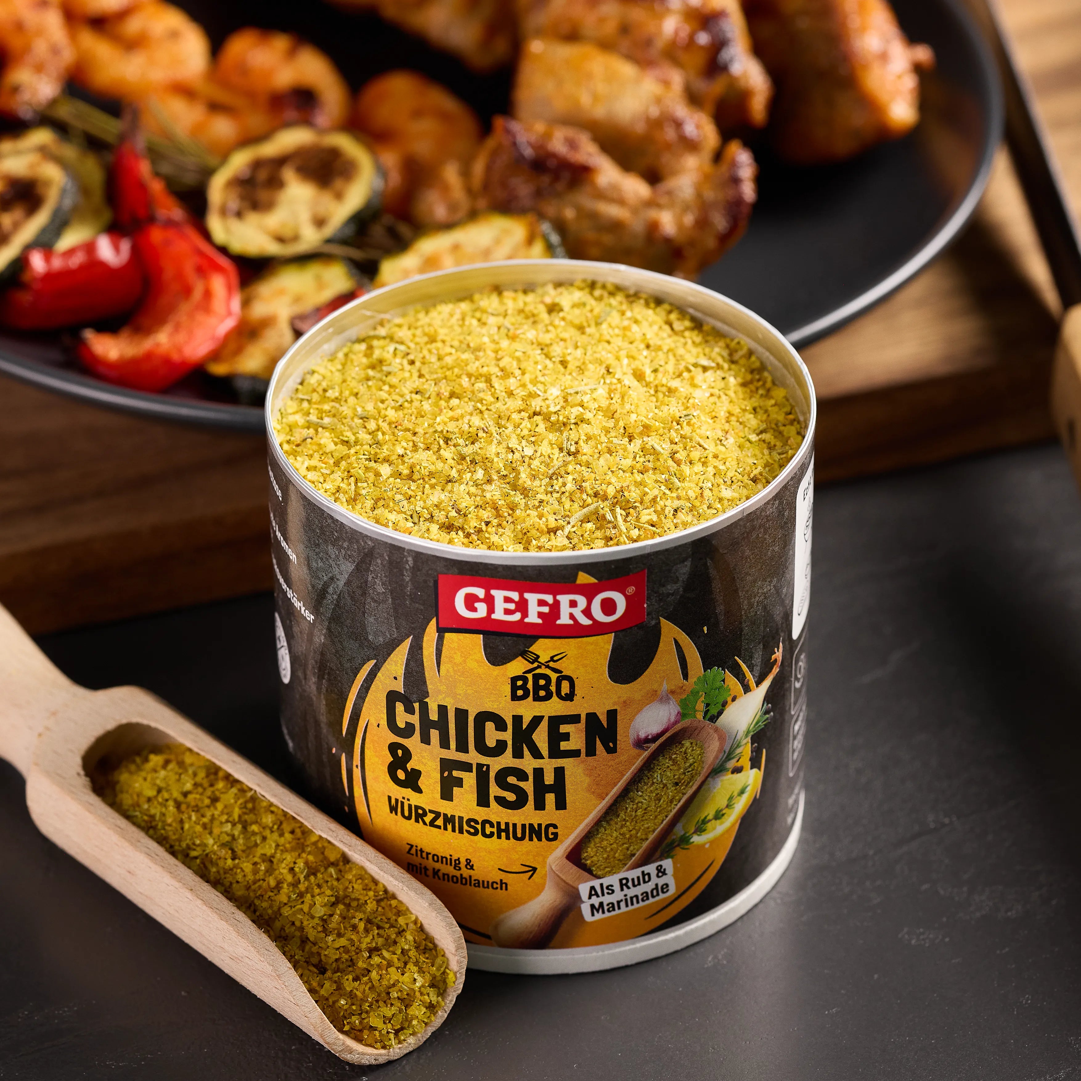 Offene Dose GEFRO BBQ Chicken & Fish Würzmischung mit Holzschaufel, vor gegrilltem Hähnchen und Gemüse.