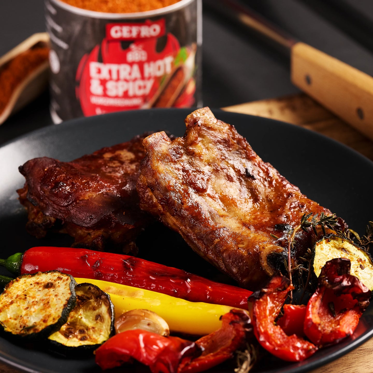 Gegrillte Spareribs auf schwarzem Teller mit Paprika und Zucchini, im Hintergrund eine Dose scharfe Gewürzmischung