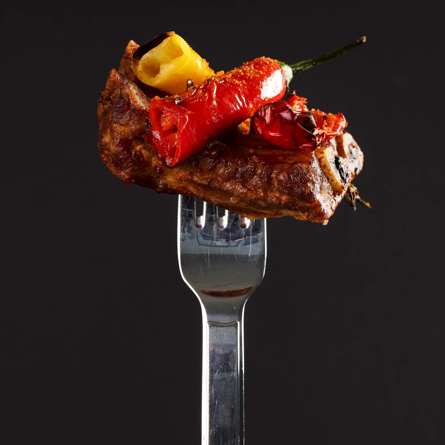 Gegrilltes Fleischstück auf einer Gabel, belegt mit roter Chili und Gemüse vor schwarzem Hintergrund