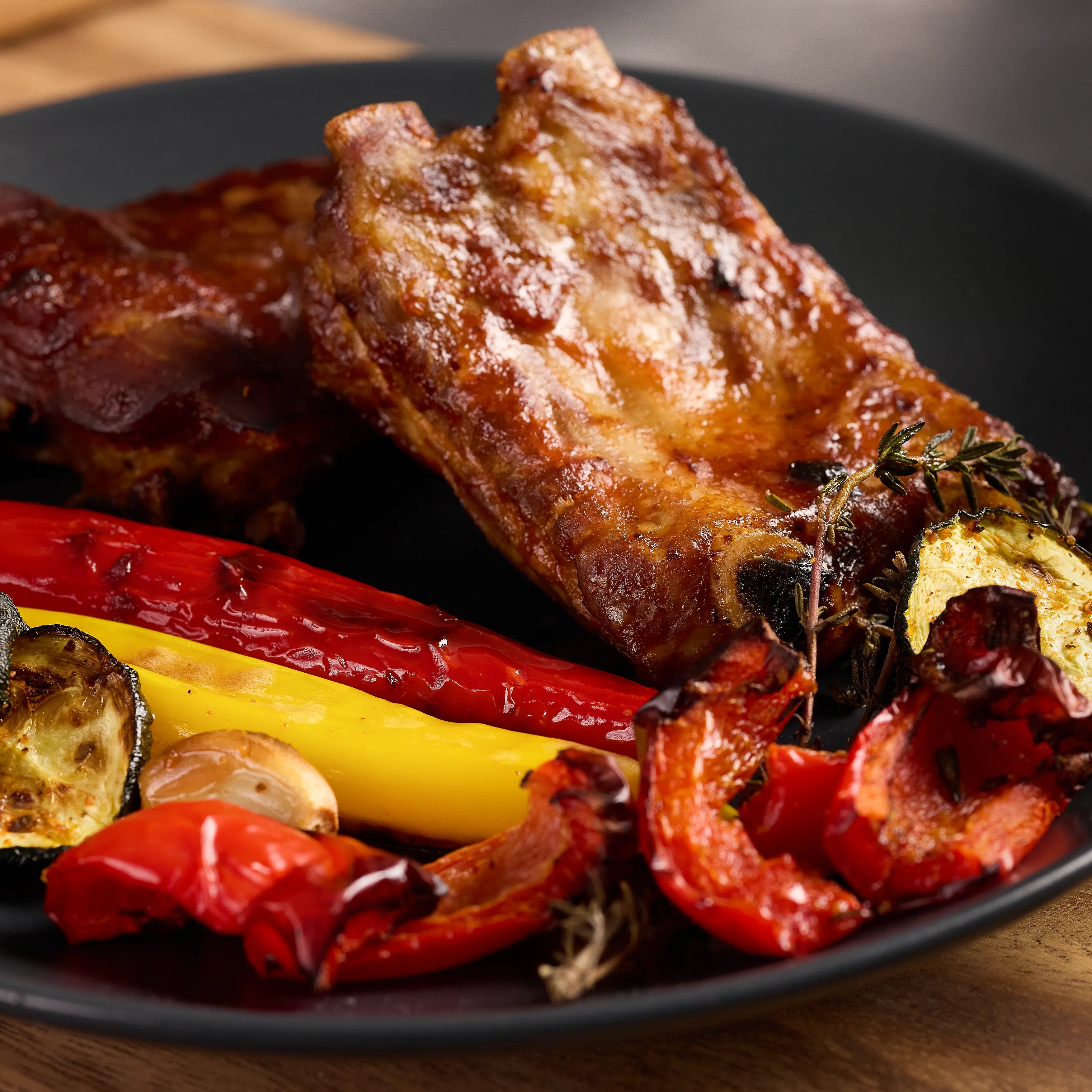 Gegrillte Schweinerippchen auf schwarzem Teller mit gerösteter Paprika, Zucchini und Knoblauch als Serviervorschlag