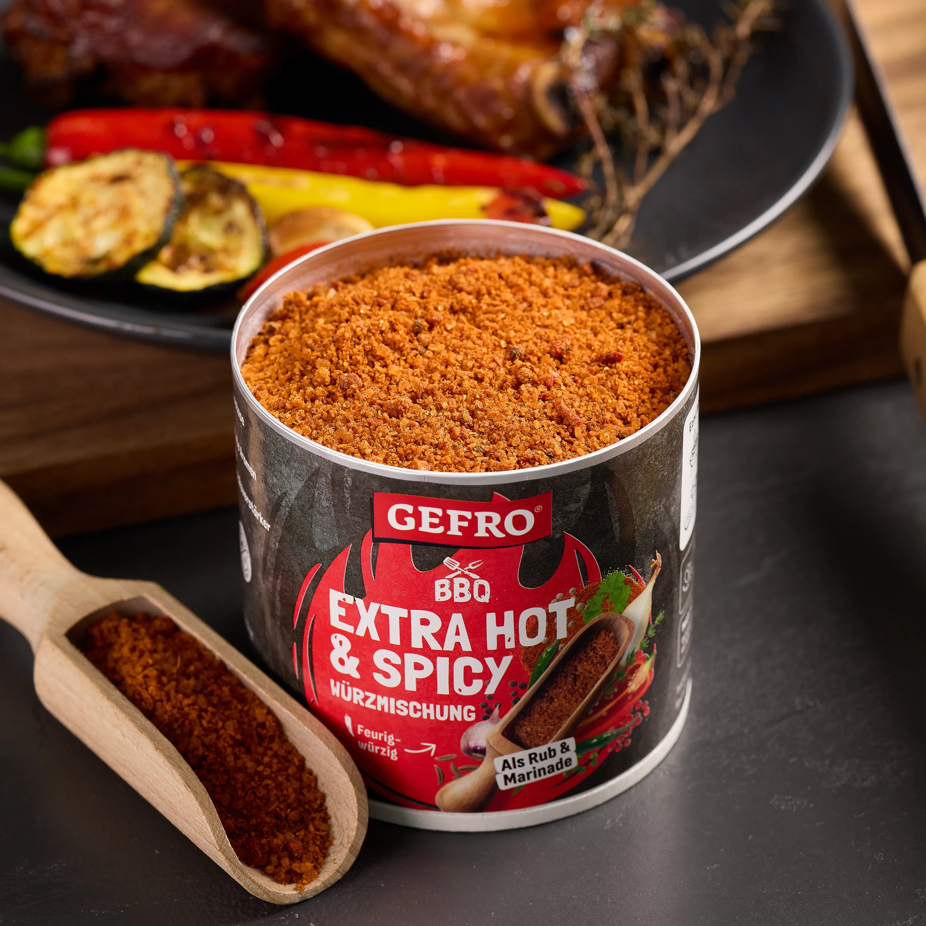 Dose GEFRO BBQ Extra Hot & Spicy Würzmischung mit Holzschaufel, vor gegrilltem Gemüse und Fleisch platziert