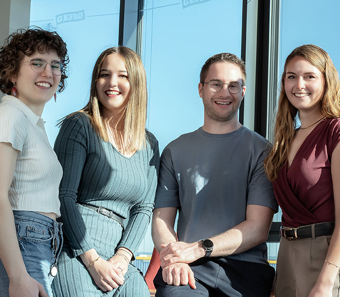 Vier junge Erwachsene stehen und sitzen lächelnd vor großen Fenstern in einem hellen Büro oder Arbeitsraum.