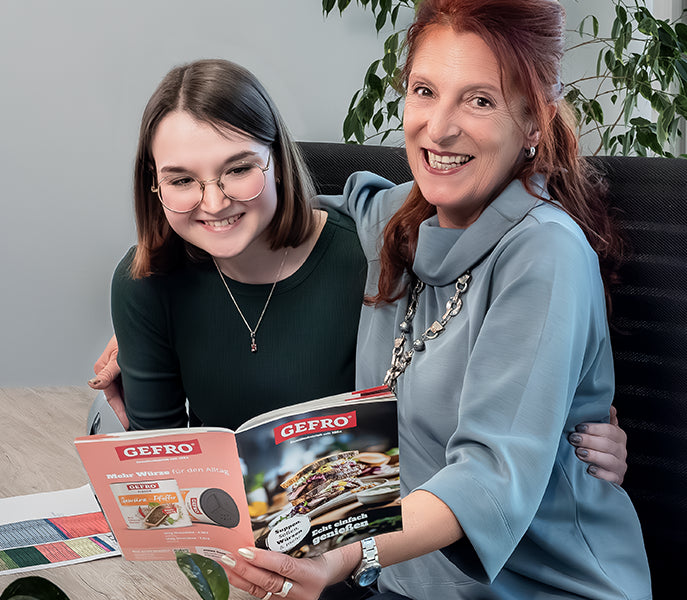 Zwei Frauen sitzen zusammen und schauen lächelnd in einen GEFRO-Katalog an einem Tisch im Büro.