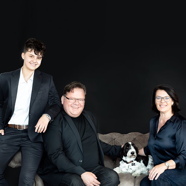 Familienporträt mit drei Personen in dunkler Kleidung und einem schwarz-weißen Hund auf einem Sofa vor schwarzem Hintergrund