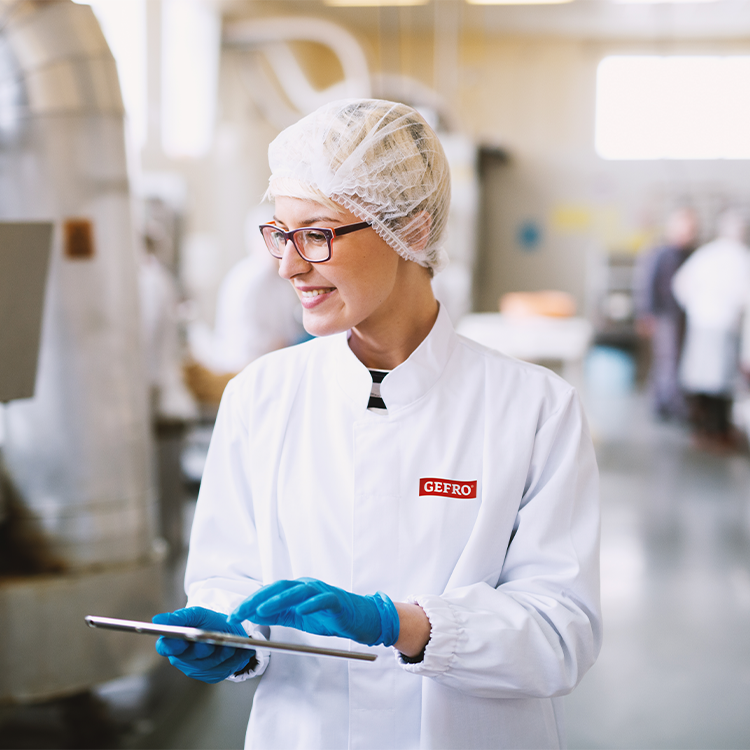 Mitarbeiterin in weißer Hygienekleidung mit Haarnetz und Handschuhen nutzt ein Tablet in der Produktion.