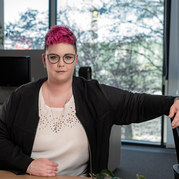 Porträt einer Frau mit pinken Haaren und Brille im Büro, sitzend am Tisch vor einem großen Fenster