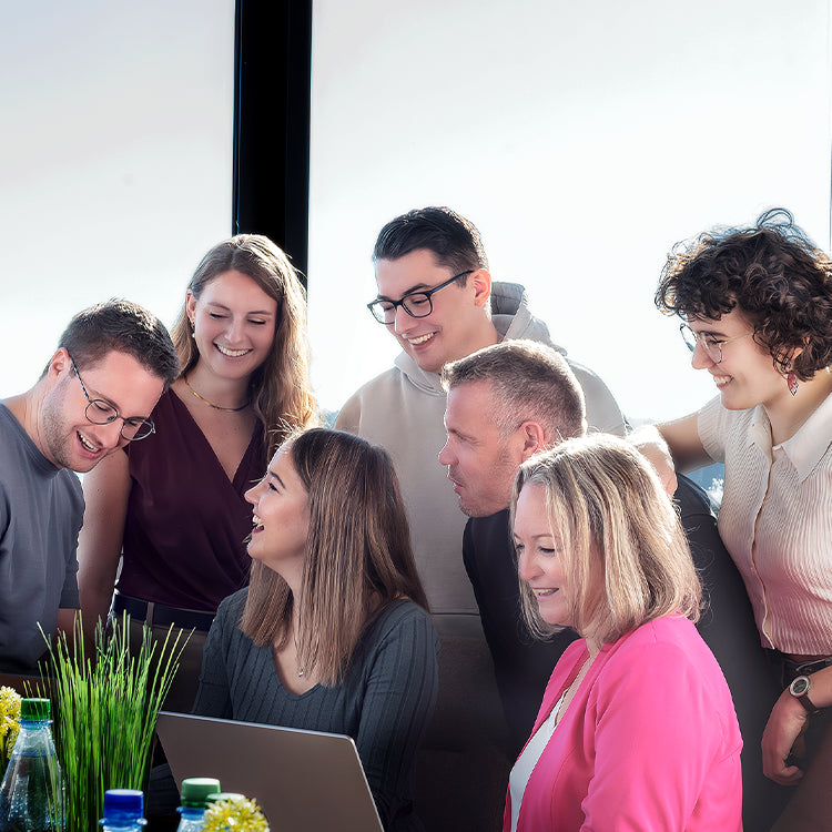 Sieben Personen schauen gemeinsam auf einen Laptop im hellen Büro und besprechen sich in freundlicher Stimmung