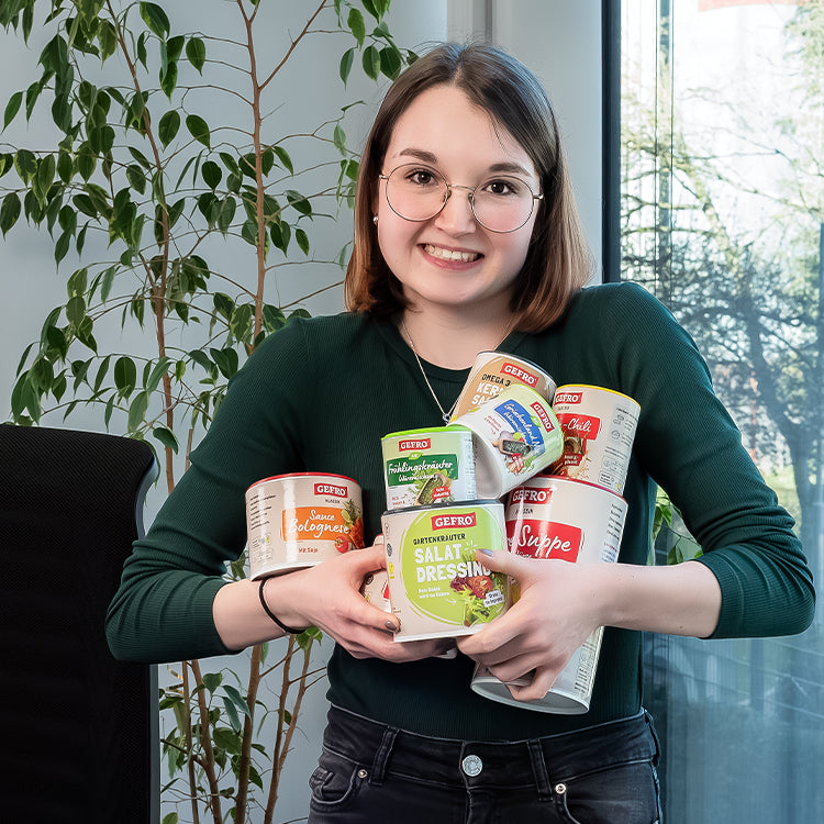 Lächelnde Frau mit Brille hält mehrere Gefro-Produkte im Arm, darunter Suppe, Salatdressing und Würzmischungen.