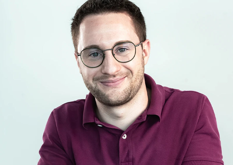 Porträt eines lächelnden Mannes mit runder Brille und bordeauxfarbenem Poloshirt vor hellem Hintergrund