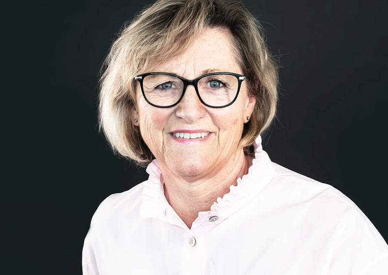 Porträt einer lächelnden Frau mit Brille und weißer Bluse vor dunklem Hintergrund, freundlich und professionell