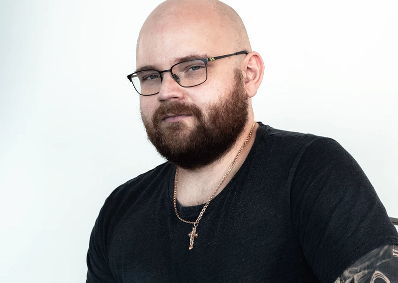 Porträt eines glatzköpfigen Mannes mit Bart, Brille, schwarzem T-Shirt und Halskette vor hellem Hintergrund