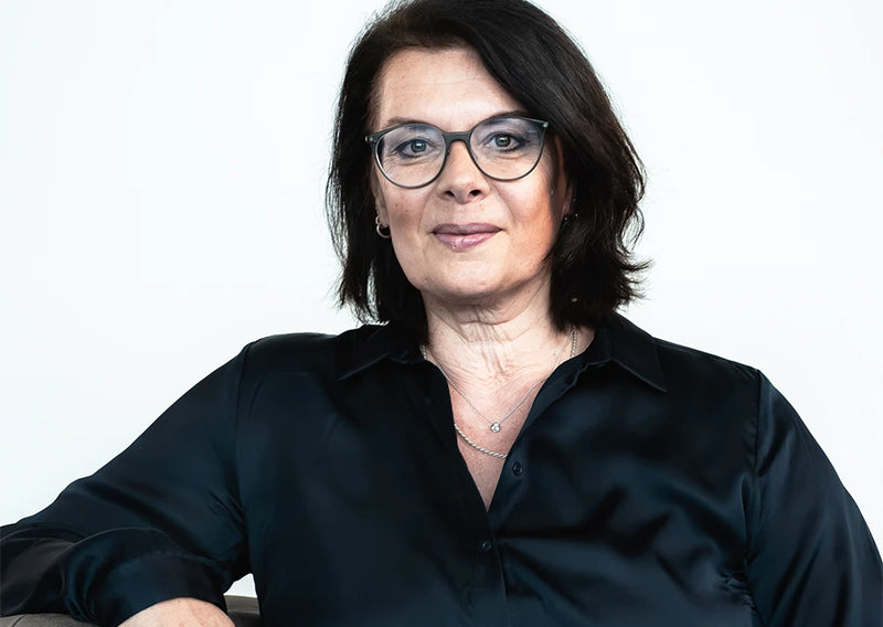 Porträt einer Frau mit Brille und schwarzer Bluse vor hellem Hintergrund, freundlich und professionell dargestellt