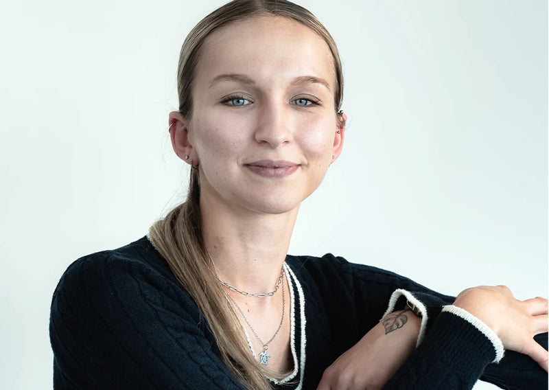 Porträt einer Frau mit Pferdeschwanz in schwarzem Pullover, mit Ketten und kleinem Tattoo am Unterarm