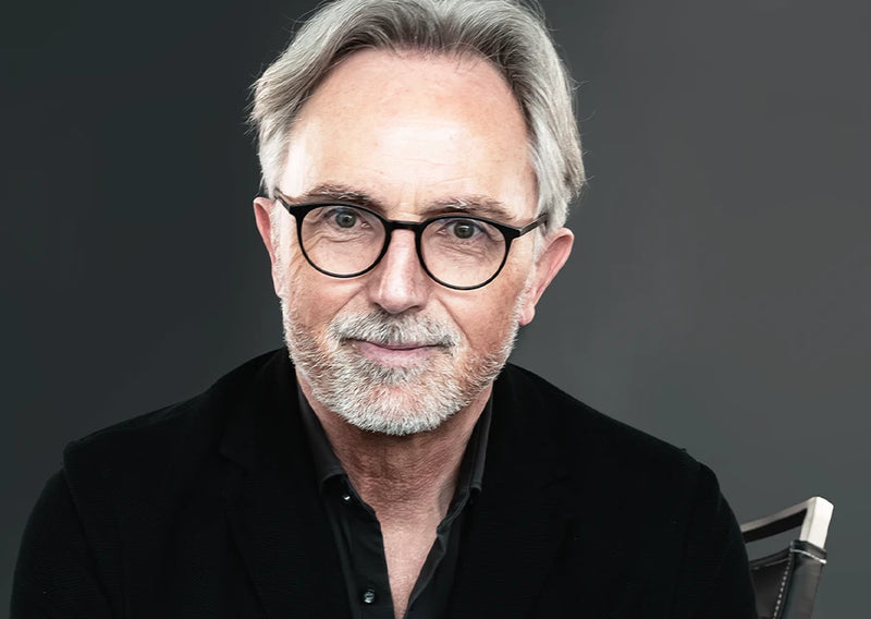 Porträt eines älteren Mannes mit grauem Haar, Brille und Bart in schwarzem Sakko vor grauem Hintergrund