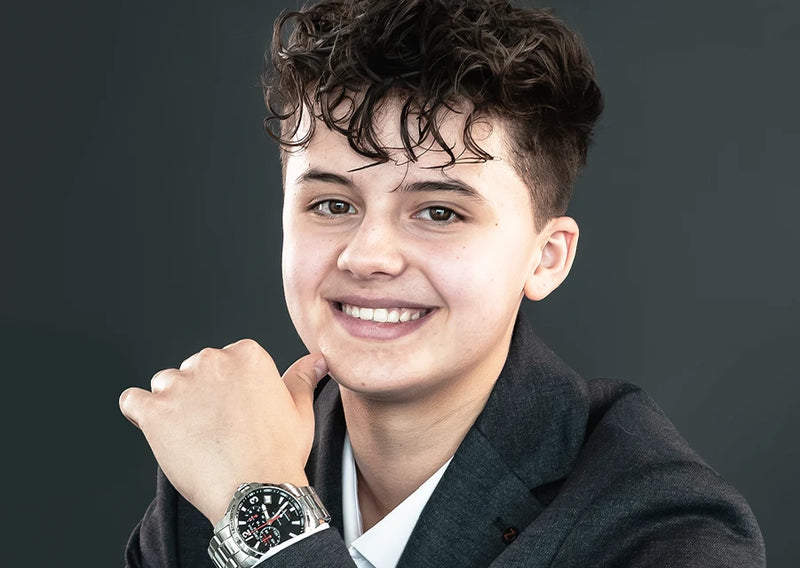 Jugendliche Person im dunklen Sakko mit Armbanduhr am Handgelenk, lächelnd vor grauem Hintergrund.