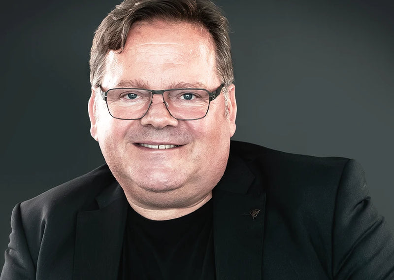 Porträt eines lächelnden Mannes mit Brille im schwarzen Sakko vor dunklem Hintergrund, frontal aufgenommen