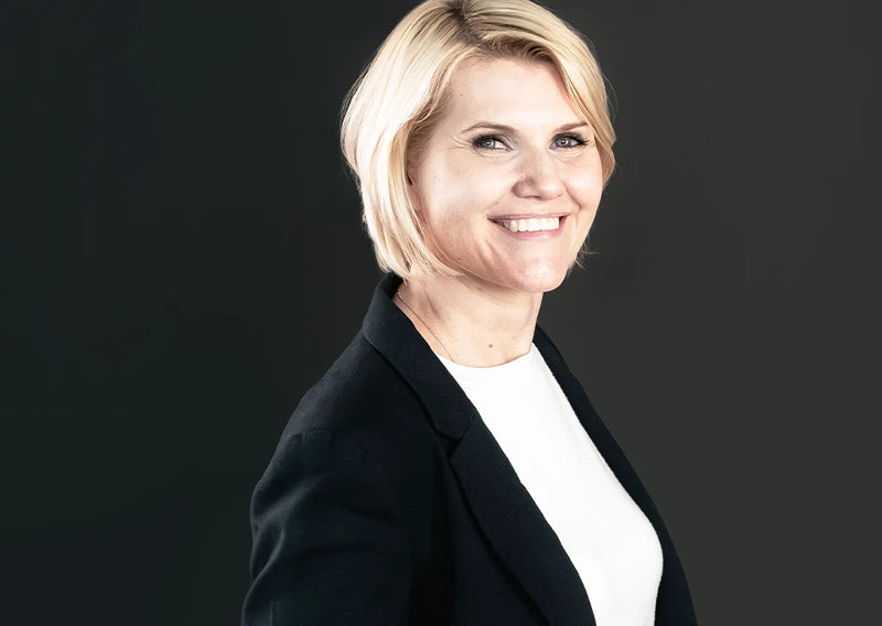 Porträt einer lächelnden Frau mit blondem Haar in schwarzem Blazer und weißem Oberteil vor dunklem Hintergrund