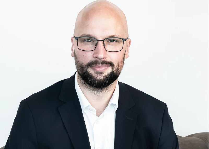Porträt eines lächelnden Mannes mit Brille, Bart, weißem Hemd und dunklem Sakko vor hellem Hintergrund