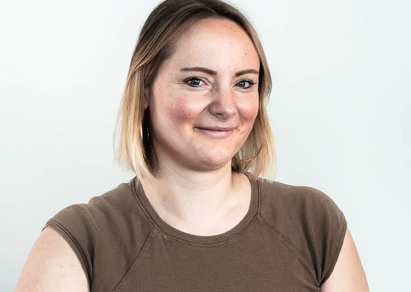 Porträt einer lächelnden Frau mit kurzen blonden Haaren, braunem Oberteil und großen Creolen vor hellem Hintergrund