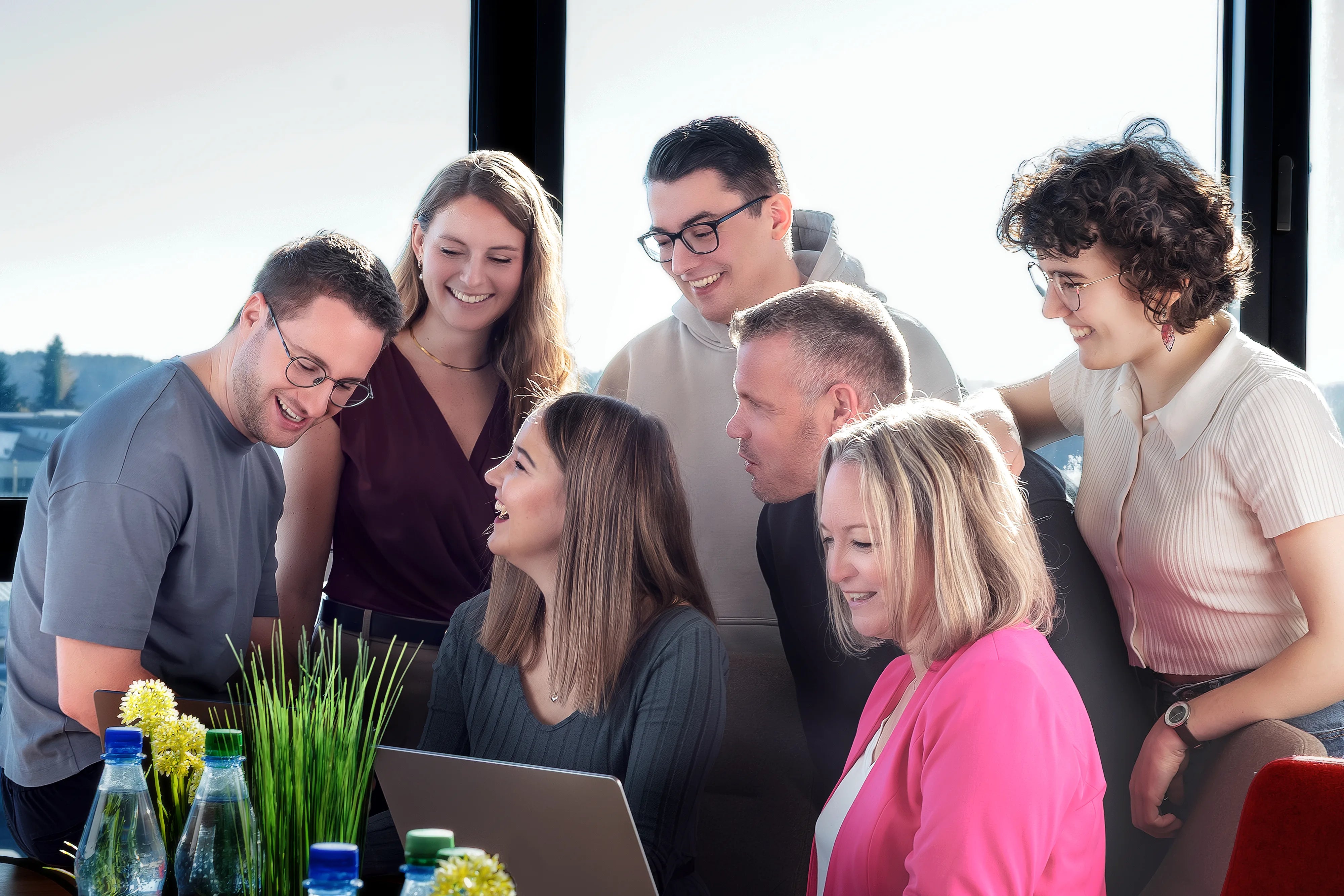 Sieben Personen schauen gemeinsam auf einen Laptop und besprechen etwas in heller Büroumgebung.
