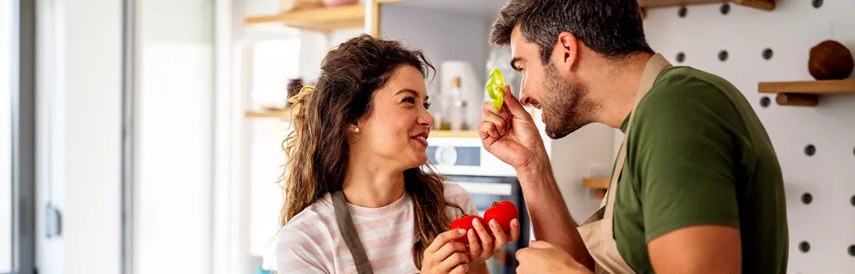 Paar mit Schürzen bereitet gemeinsam in der Küche frisches Gemüse zu und lächelt sich dabei an.