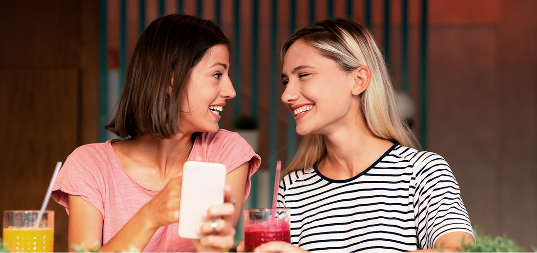 Zwei lächelnde Frauen sitzen mit Smartphone und Getränken im Café und schauen sich freundlich an.