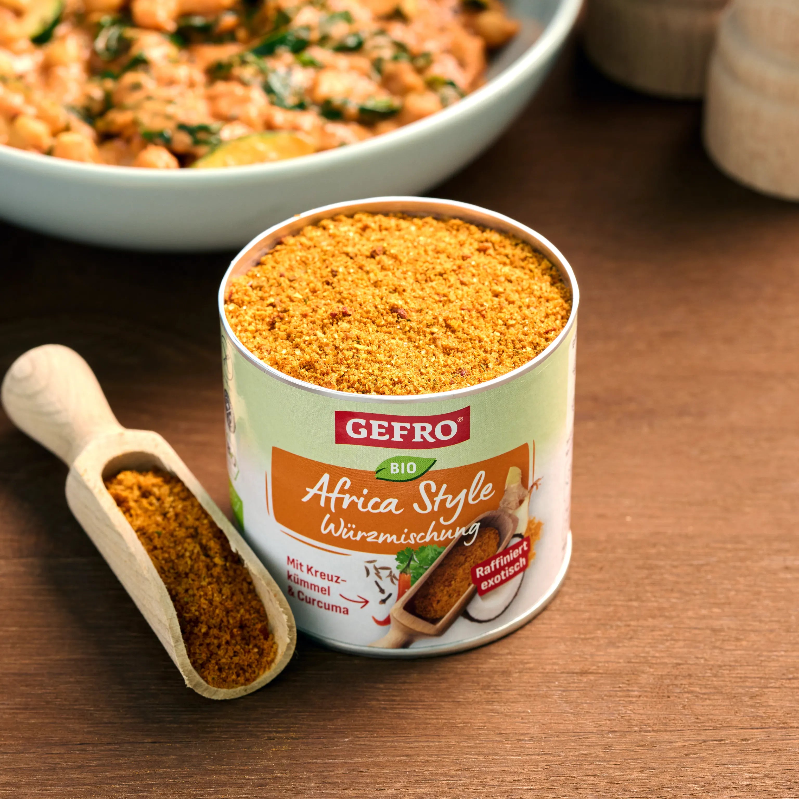 Offene Dose GEFRO Bio Africa Style Würzmischung mit Holzschaufel auf Tisch, im Hintergrund ein zubereitetes Gericht