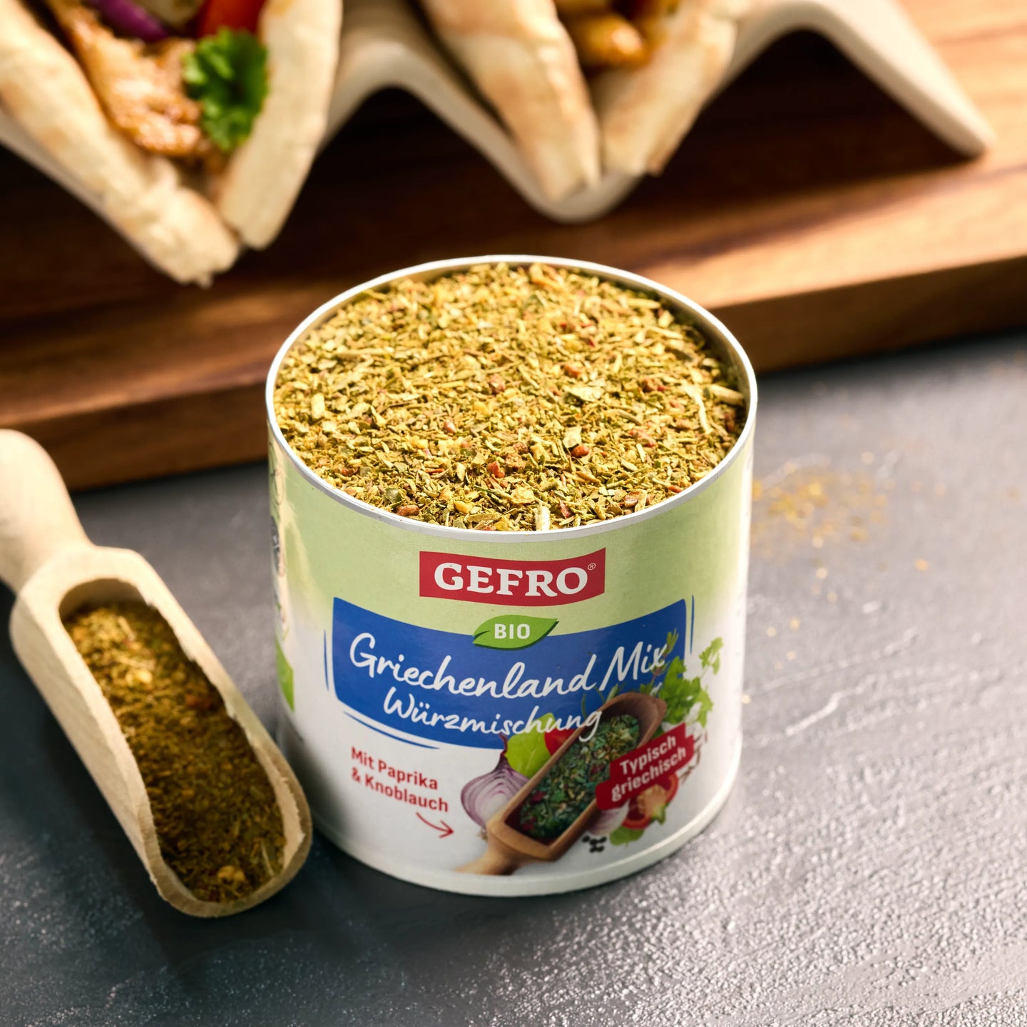 Offene Dose GEFRO Bio Griechenland-Mix Würzmischung mit Kräutern, daneben Holzschaufel auf dunkler Fläche