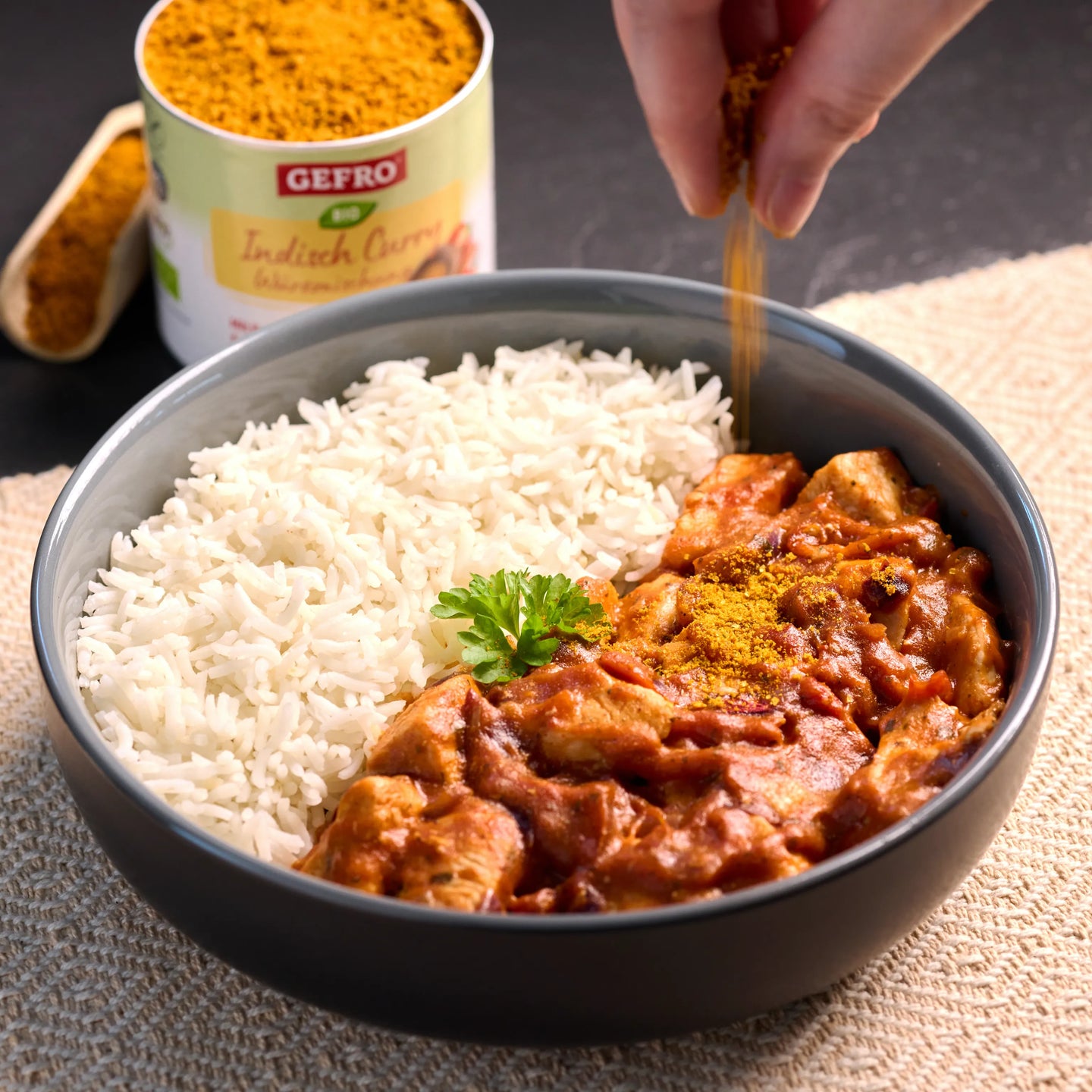 Schüssel mit Reis und cremigem Curry, darüber wird Gewürz gestreut; im Hintergrund steht eine Dose Currypulver.