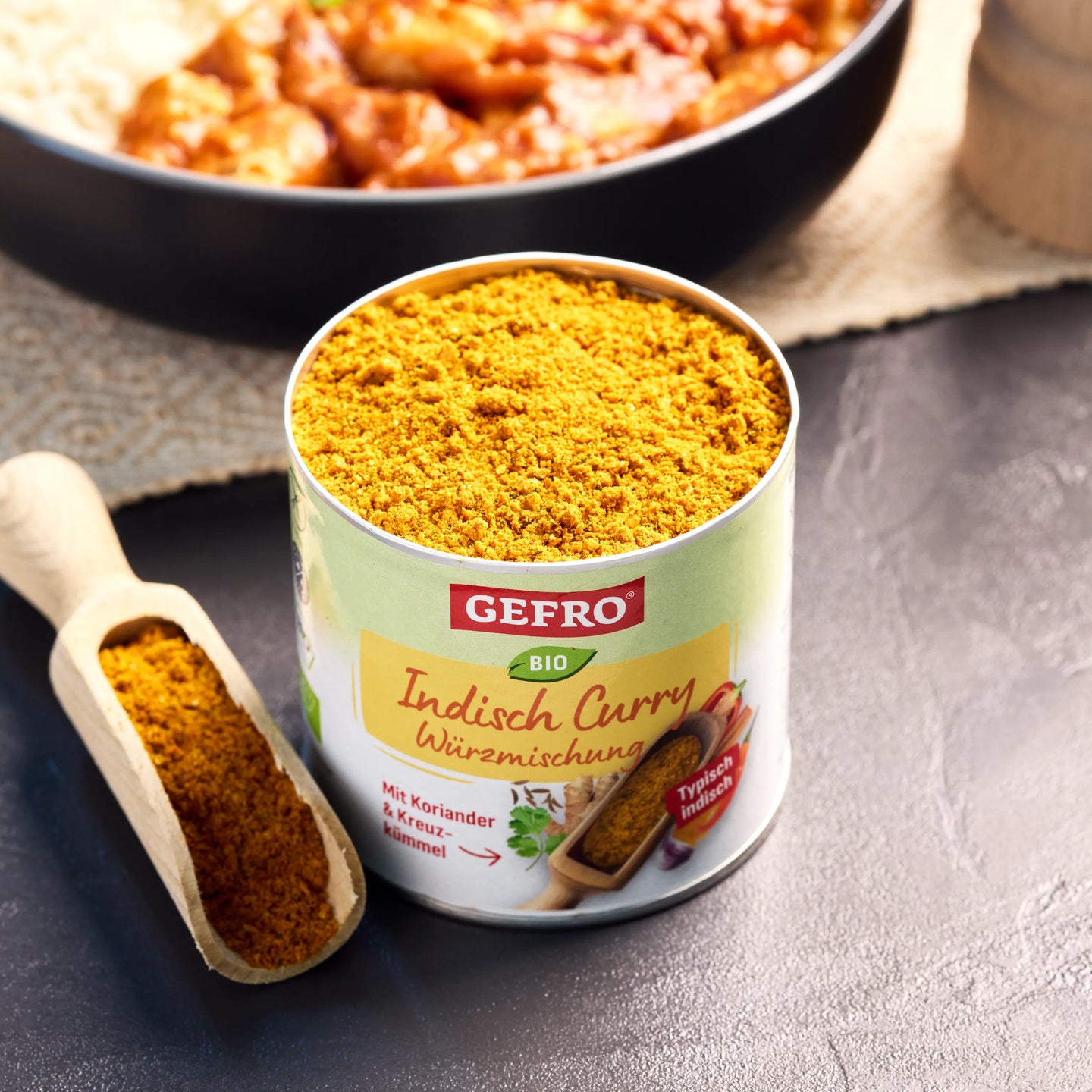 Offene Dose mit Bio Indisch Curry Würzmischung von Gefro, daneben Holzschaufel mit Gewürz und Gericht im Hintergrund