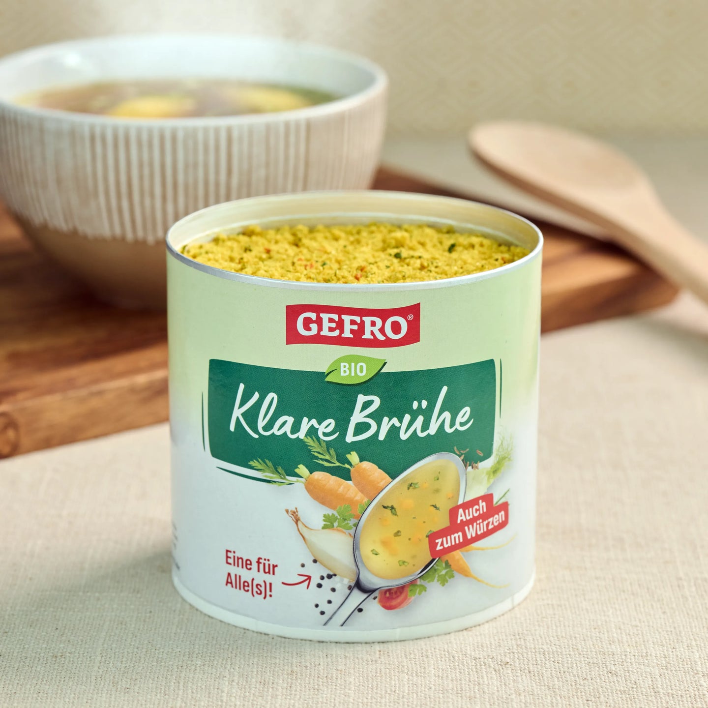 Dose GEFRO Bio Klare Brühe mit geöffnetem Inhalt auf Tisch, dahinter eine Schüssel Suppe und ein Holzlöffel