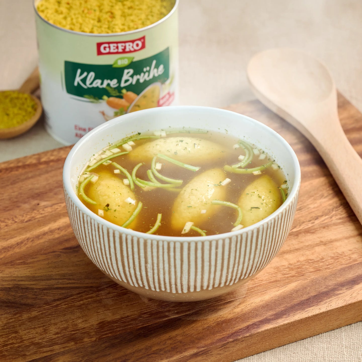 Schüssel mit klarer Brühe und kleinen Klößen auf Holzbrett, dahinter Dose Gefro Klare Brühe und Holzlöffel