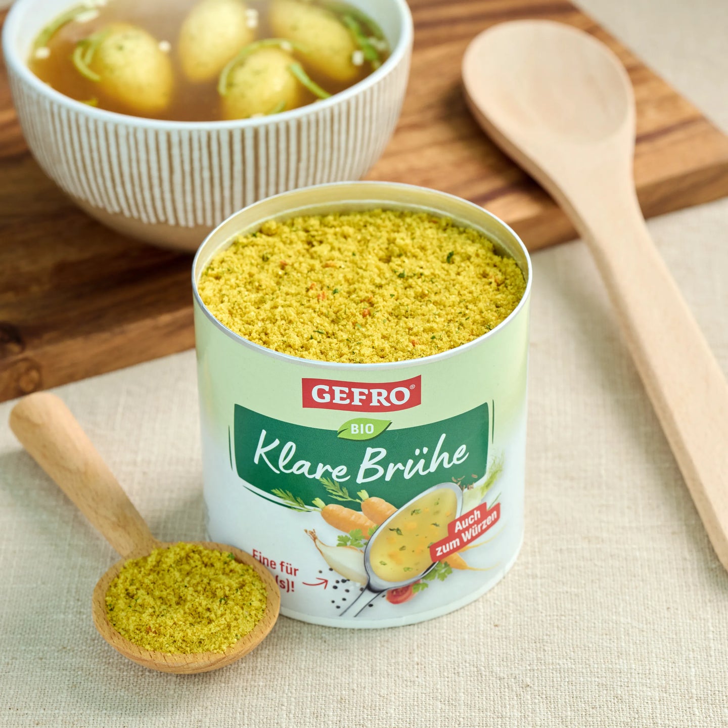 Offene Dose GEFRO Bio Klare Brühe mit Pulverlöffel, daneben eine Schüssel Brühe auf Holzbrett in der Küche
