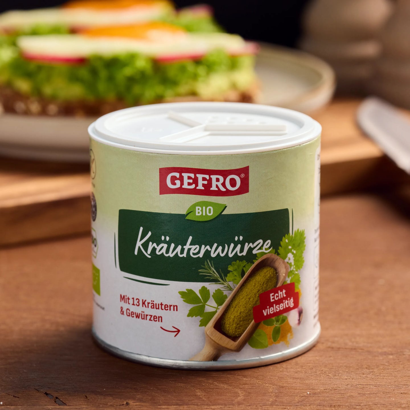 Dose GEFRÖ Bio Kräuterwürze mit weißem Deckel auf Holztisch, im Hintergrund ein belegtes Brot unscharf sichtbar