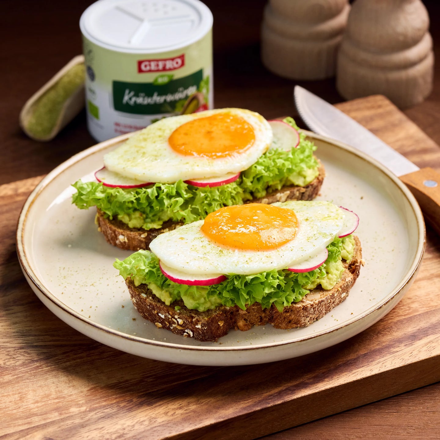 Zwei belegte Brote mit Avocado, Salat, Radieschen und Spiegelei auf einem Teller, daneben Gewürzdose und Messer
