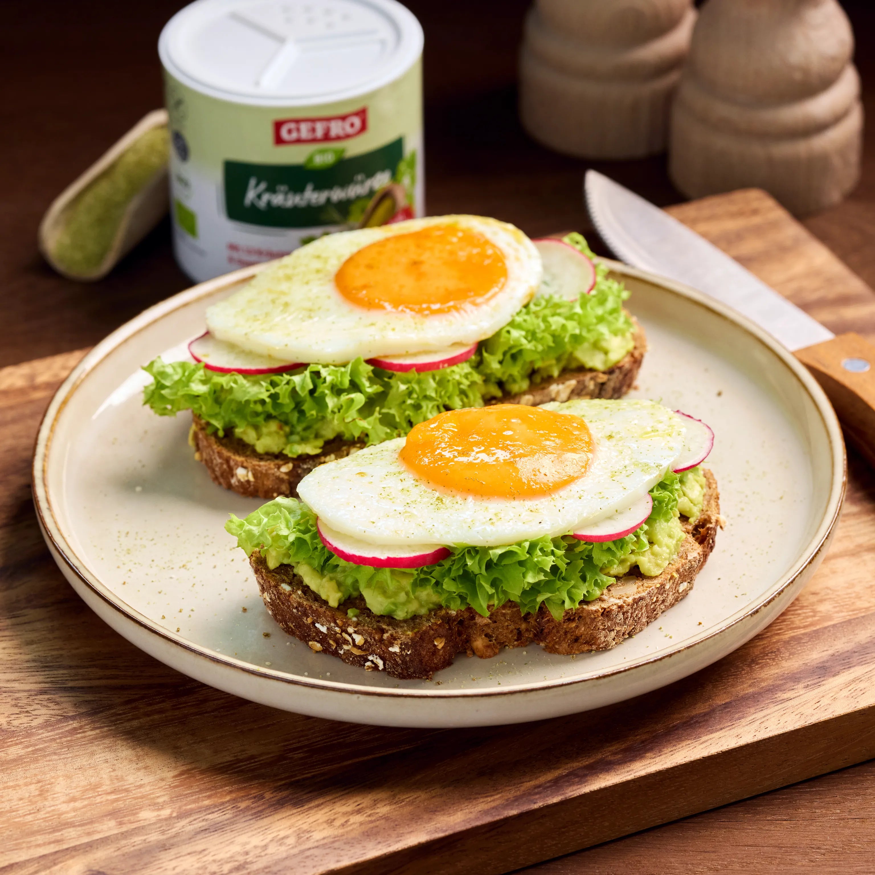 Zwei belegte Brote mit Avocado, Salat, Radieschen und Spiegelei auf einem Teller, daneben Gewürzdose und Messer