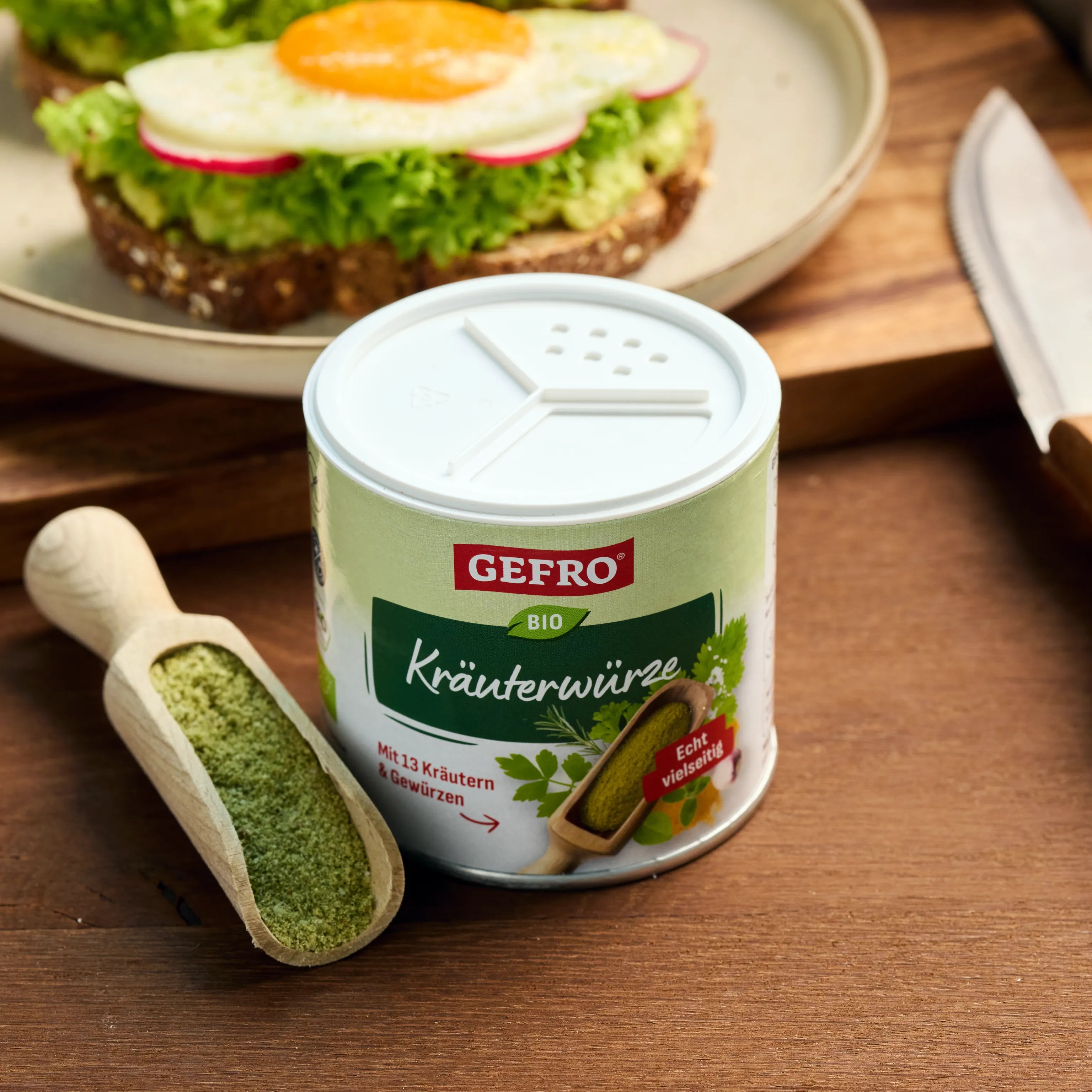 Dose GEFRO Bio Kräuterwürze mit Holzschaufel auf Tisch, dahinter belegtes Brot mit Salat und Spiegelei.
