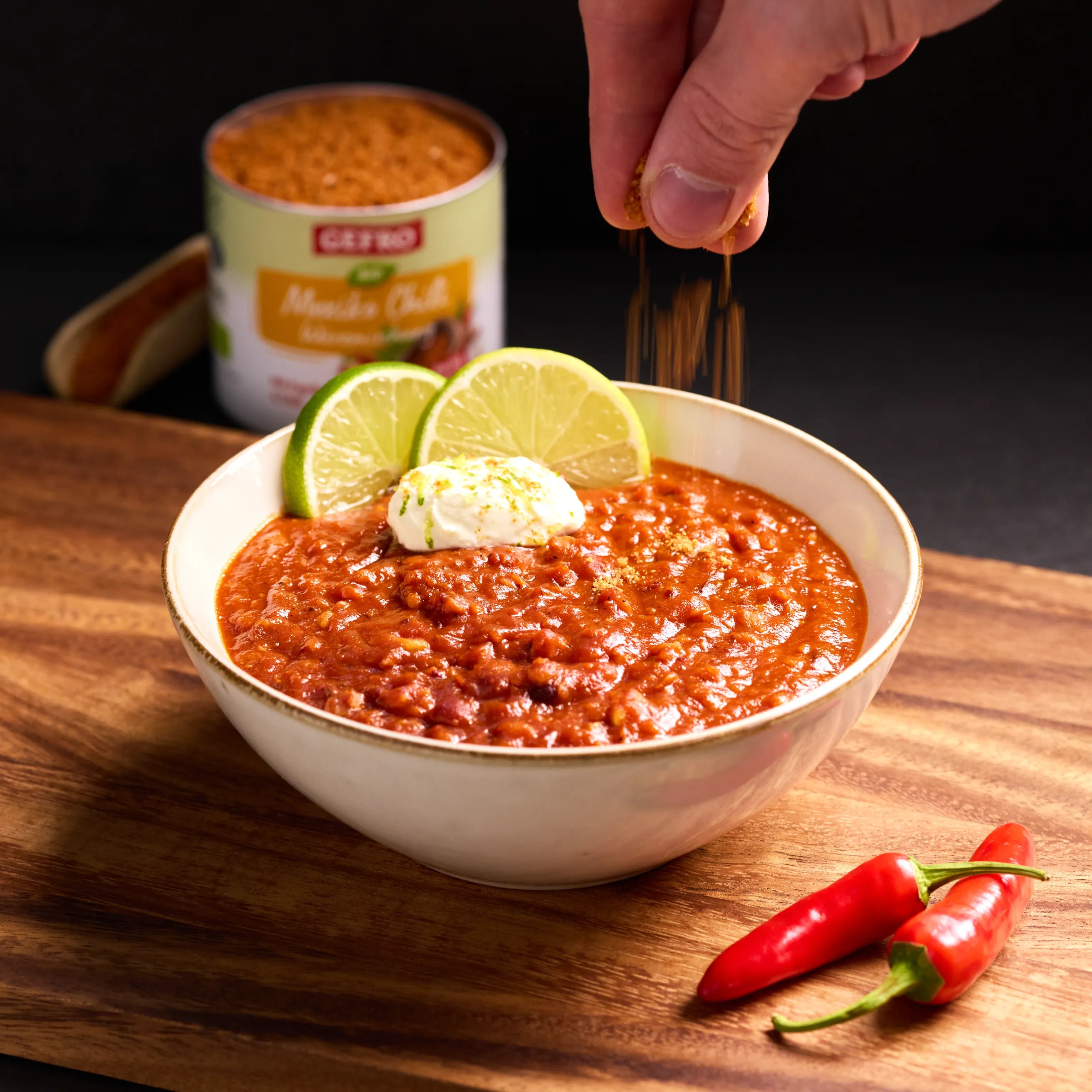 Schüssel mit rotem Chili, garniert mit Limette und Creme; eine Hand streut Gewürz darüber, zwei Chilis daneben.