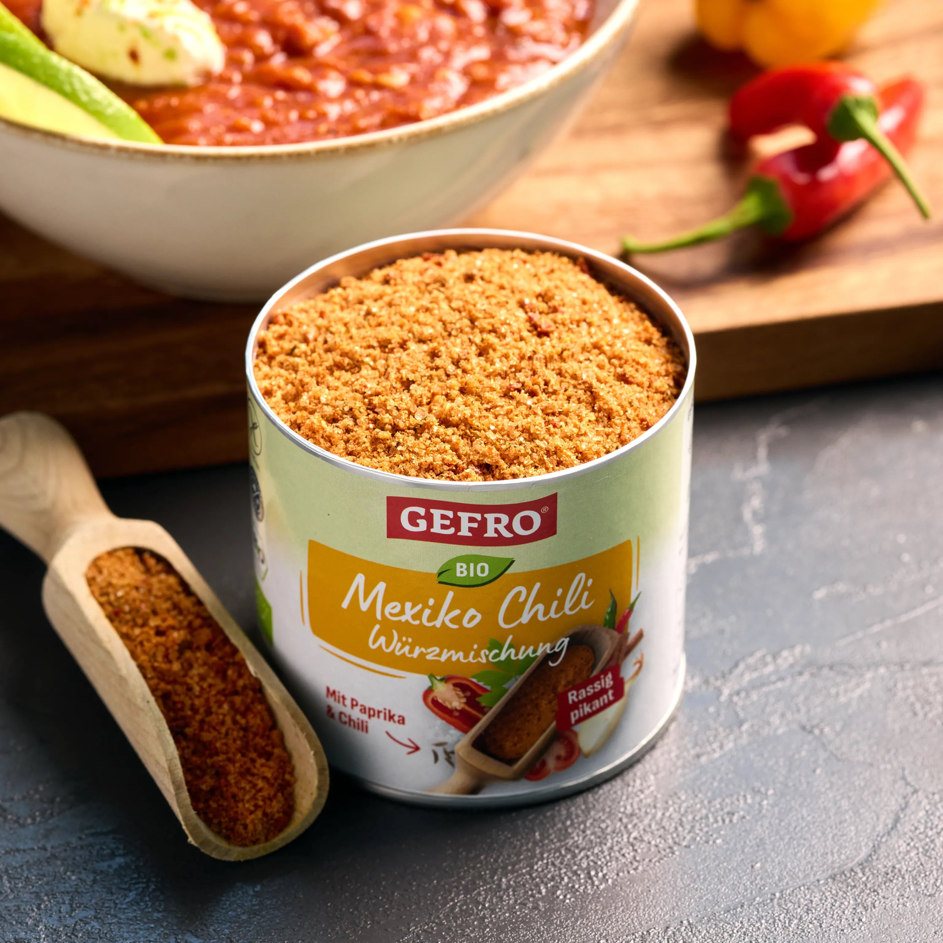Offene Dose GEFRO Bio Mexiko Chili Würzmischung mit Gewürzschaufel, davor Chili-Gericht und frische Peperoni.