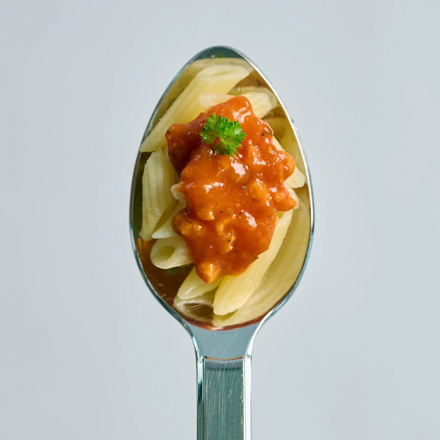 Löffel mit Penne und roter Tomatensoße, garniert mit etwas Petersilie, vor hellem Hintergrund in Nahaufnahme