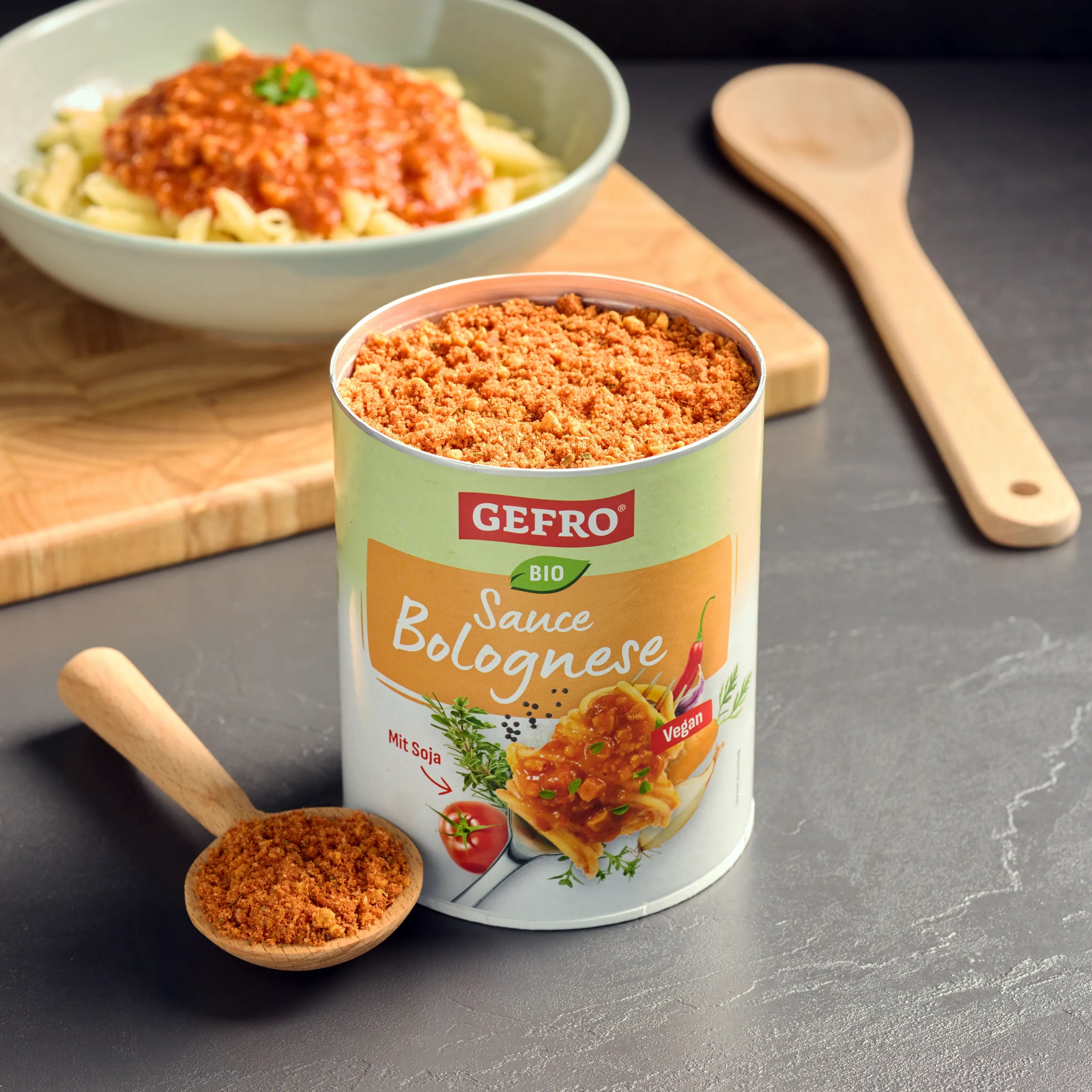 Dose mit GEFRO Bio Sauce Bolognese Pulver und Holzlöffel davor, im Hintergrund ein Teller mit Pasta und roter Sauce