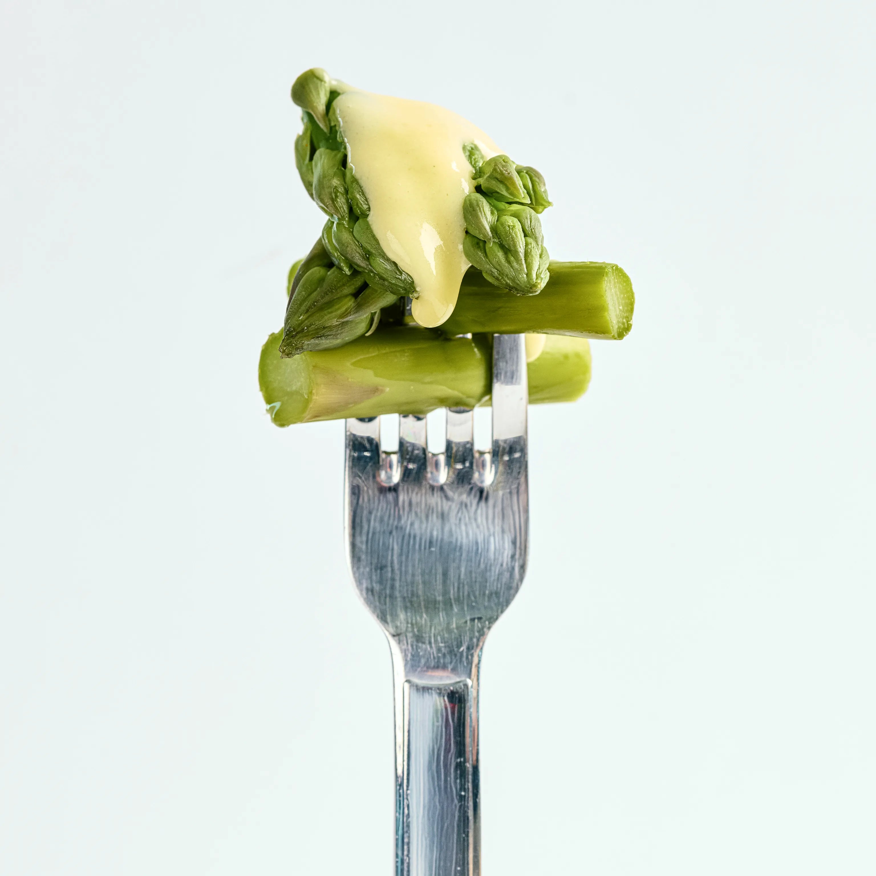 Grüne Spargelstücke auf einer Gabel mit heller Sauce, nah aufgenommen vor schlichtem, hellem Hintergrund