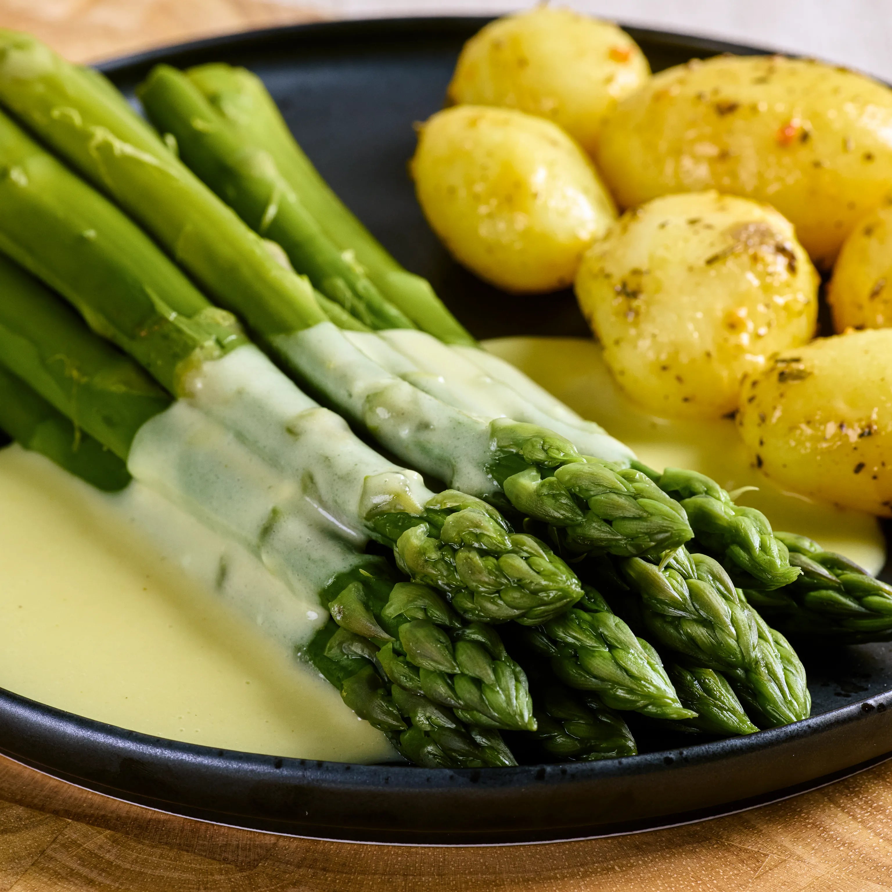 Grüner Spargel mit heller Sauce und kleinen gewürzten Kartoffeln auf schwarzem Teller als servierte Mahlzeit