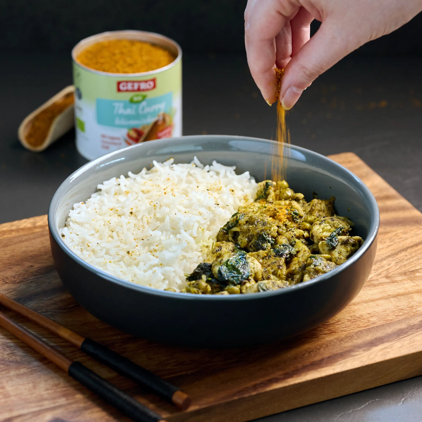 Schale mit Reis und Curry, darüber wird Gewürz gestreut; im Hintergrund steht eine Dose Thai-Curry-Gewürz.