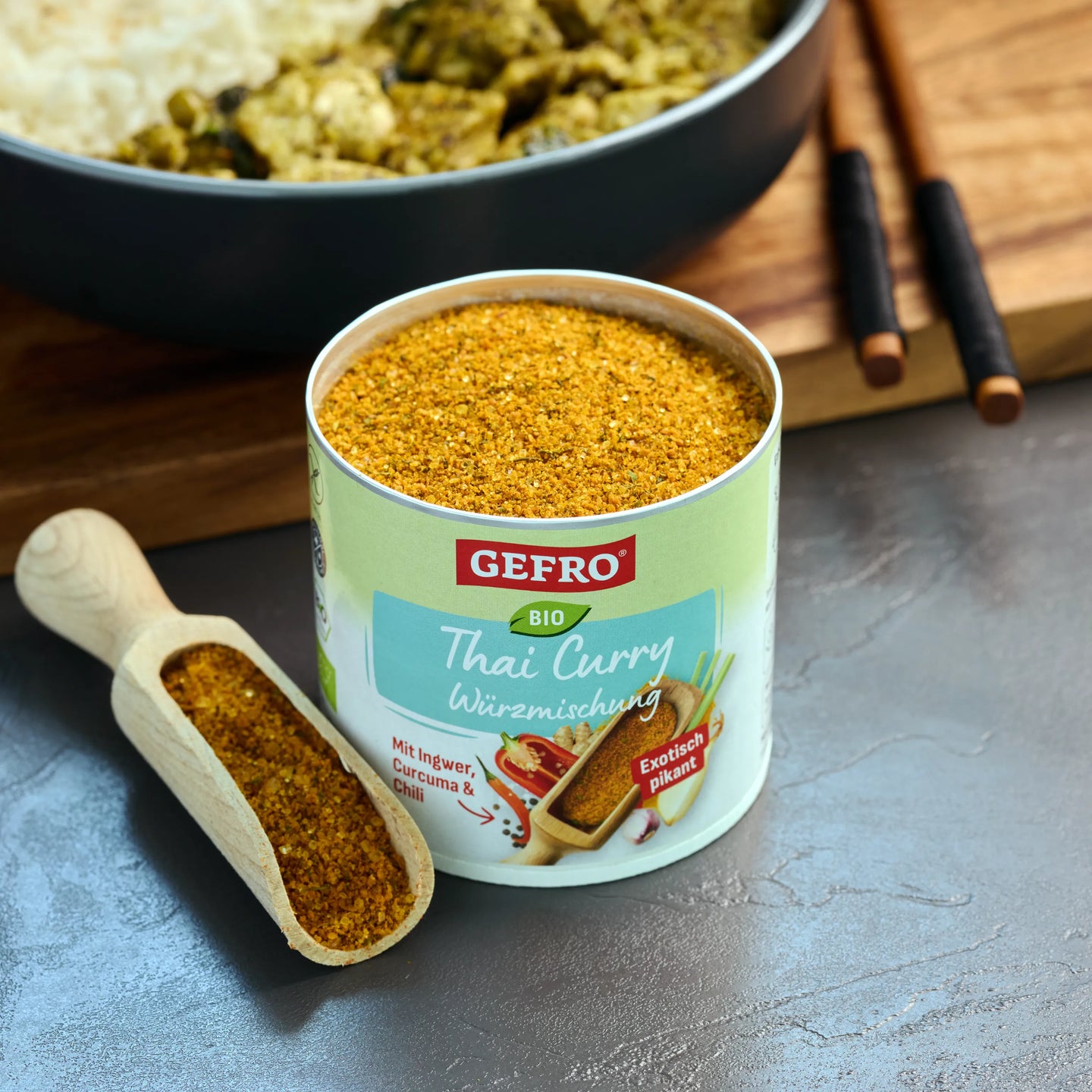Offene Dose GEFRO Bio Thai Curry Würzmischung mit Holzschaufel voller Gewürz, vor einem angerichteten Currygericht