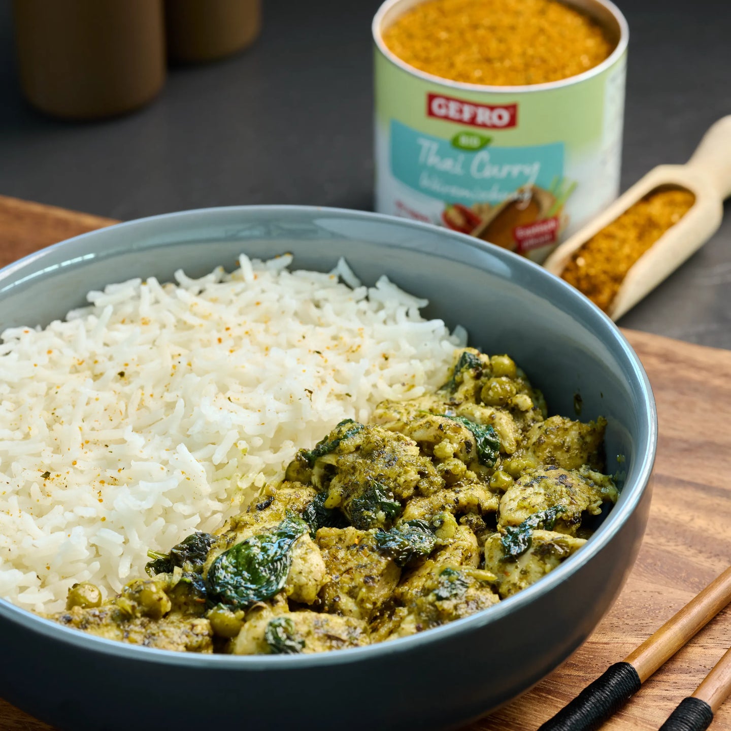Schüssel mit weißem Reis und grünem Thai-Curry mit Gemüse, dahinter Dose Gewürzmischung und Holzlöffel.